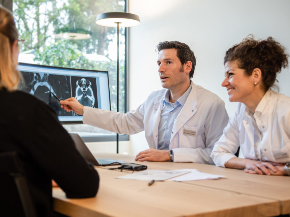 Ärzte besprechen MRI Bilder mit Patientin am Holztisch im Sprechstundenzimmer